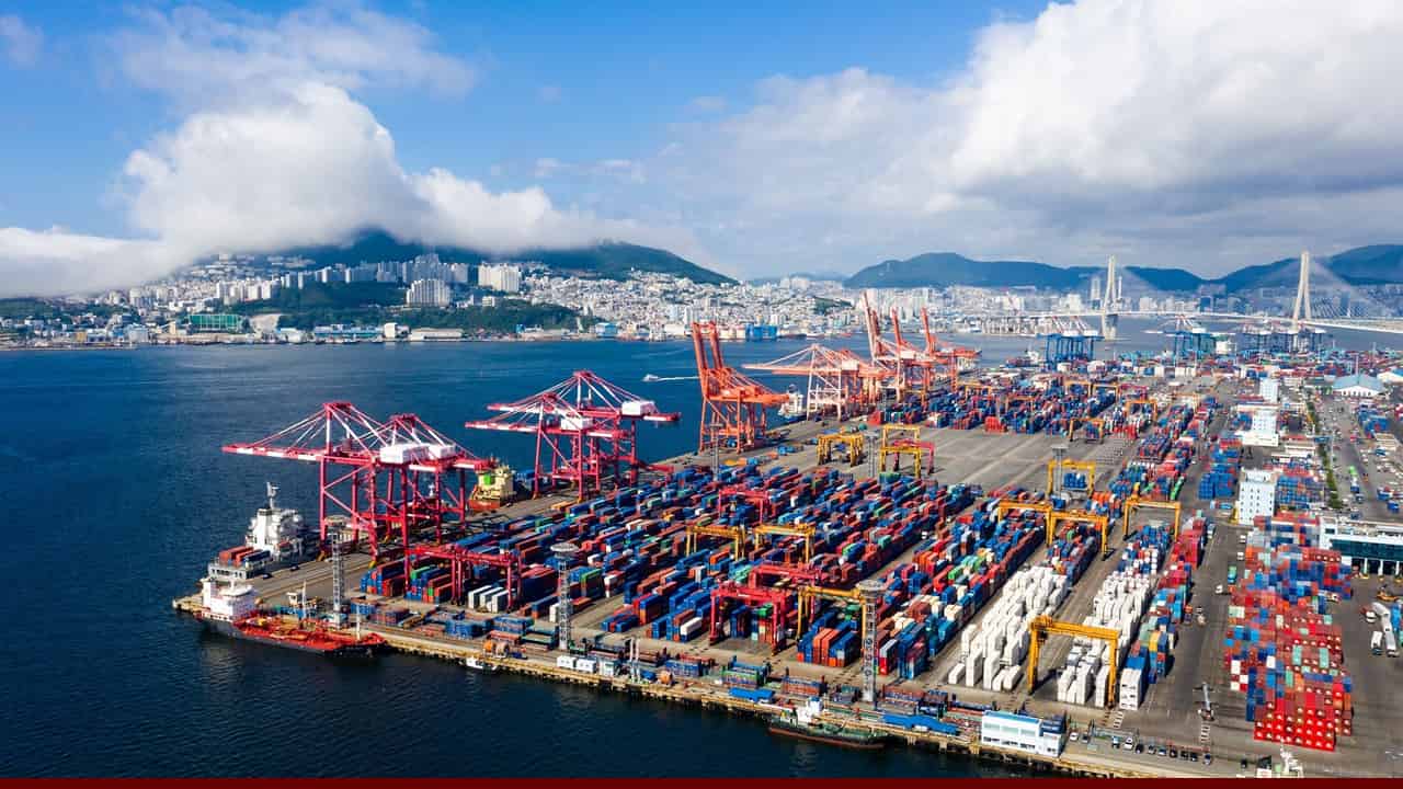 Container port packed with colorful shipping containers and cranes along a coastal city skyline, illustrating global trade and rising logistics pressure.