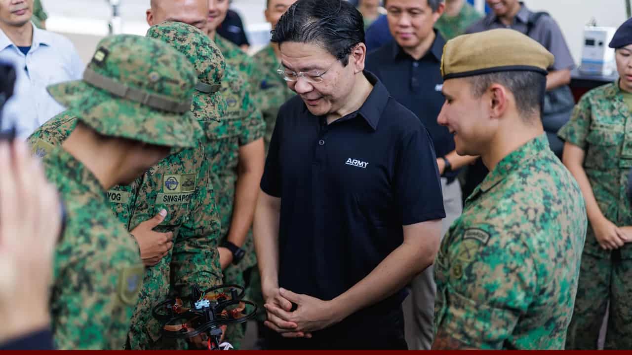 A group of soldiers in camouflage uniforms gather around a man in a black shirt as they examine a small drone, with the man observing closely while others look on.