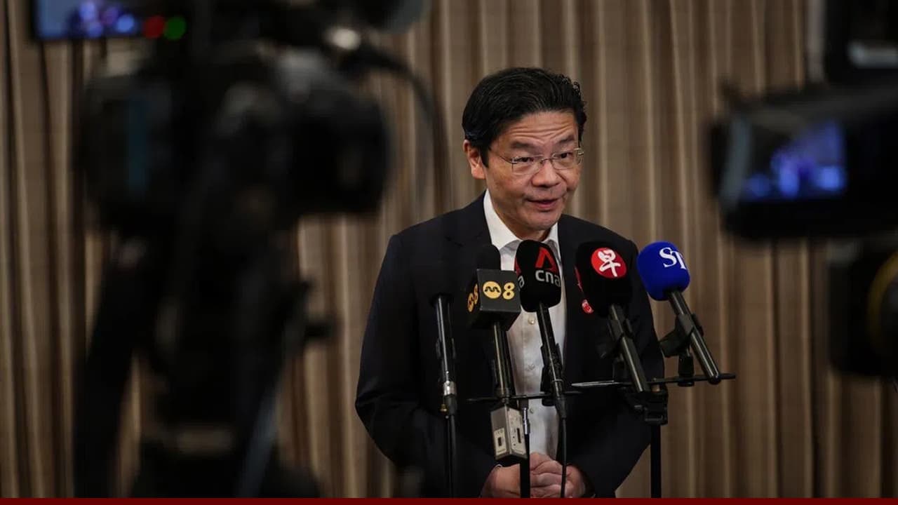 Lawrence Wong speaking at a press conference, standing behind multiple microphones with media logos, addressing reporters indoors.